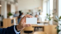 Businessman holding blank white business card in modern office environment with colleagues and plants in background.