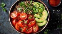 Fresh quinoa bowl with sliced avocado, cherry tomatoes, and herbs served on dark background with red spices and sesame seeds for healthy lunch menu.