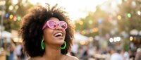 Happy young African American woman with curly afro hair wearing pink sunglasses and green earrings laughing joyfully at outdoor festival or event.