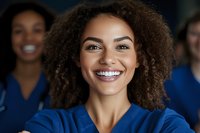 Young African American female healthcare worker with natural curly hair and bright smile wearing blue medical scrubs, with colleagues in background.