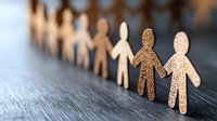 Paper cutout figures holding hands in chain formation on dark wooden surface, symbolizing unity, diversity, community and social connection. Shallow depth of field.