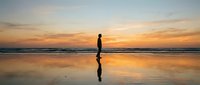 Surfer holding board standing on wet beach sand at golden sunset, reflected in water. Coastal lifestyle and water sports.