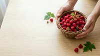 Fresh raspberries in wicker basket held by hands on wooden table with scattered berries and green leaves, suitable for food and recipe content.