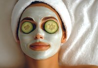 Young woman with clay facial mask and cucumber slices on eyes relaxing during spa treatment. White towel wrapped around head, skincare routine.