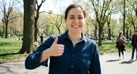 Happy young Caucasian woman giving thumbs up gesture in sunny city park during spring day. Positive female in denim shirt smiling outdoors for success concept.