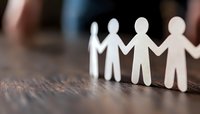 Paper cutout figures holding hands in chain on wooden surface, symbolizing unity, teamwork, and community support. Shallow depth of field creates artistic blur.