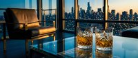 Luxury penthouse interior with whiskey glasses overlooking illuminated city skyline at dusk through floor-to-ceiling windows creating elegant urban lifestyle ambiance.