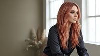 Professional young Caucasian businesswoman with long wavy red hair in black blazer sitting thoughtfully by office window during daytime meeting preparation.
