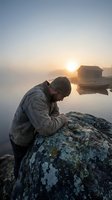 Man in beanie sitting on lichen-covered rock by misty lake at sunrise with wooden boathouse reflecting in calm water for outdoor lifestyle content.