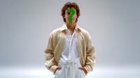 Young Caucasian man in casual beige jacket and white shirt with green question mark symbol over face standing against gray background, expressing uncertainty concept.