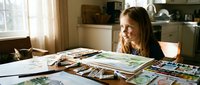 Young Caucasian girl organizing family photos and memories at kitchen table, creating scrapbook album with nostalgic vintage photographs in warm sunlit home interior.