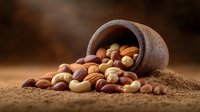 Mixed nuts spilling from rustic clay pot on earthy background, featuring cashews, almonds and hazelnuts in warm natural lighting, creating appetizing composition.