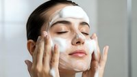 Young woman applying white facial cleanser with closed eyes in bathroom for skincare routine and beauty treatment.