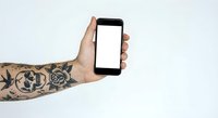 Tattooed hand holding smartphone with blank white screen against white background for mockup and app presentation purposes.