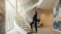 Professional businesswoman in black suit standing on modern glass staircase in luxury office building with natural light streaming through windows.
