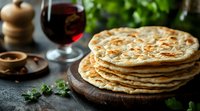 Stack of fresh homemade flatbread on wooden board with red wine glass and herbs, rustic food photography with moody lighting and dark background.
