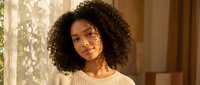Young African American woman with curly hair in cozy sweater enjoying warm sunlight through window at home creating peaceful morning atmosphere.