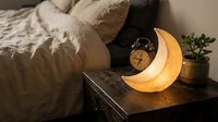 Cozy bedroom nightstand with glowing crescent moon lamp, alarm clock and small plant creating warm ambient lighting for relaxation and sleep.