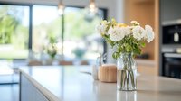 Fresh white peonies in glass vase on marble kitchen counter, bright modern interior with natural light from large windows and blurred background.