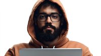 Young Middle Eastern man with beard and glasses wearing brown hoodie working on laptop computer against white background, modern freelance lifestyle concept.