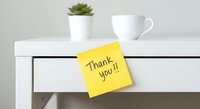 Yellow sticky note with handwritten thank you message on white desk drawer with coffee cup and succulent plant in modern minimalist workspace setting.