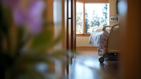 Empty hospital room with adjustable bed seen through doorway, soft natural lighting from window creates peaceful atmosphere. Medical facility interior.