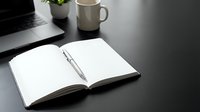 Open notebook with pen on dark desk beside laptop and coffee mug in modern office workspace for business planning and productivity.