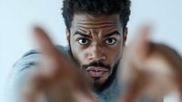 Young African American man with beard and curly hair expressing surprise with raised eyebrows and open mouth, showing emotional reaction against light background.