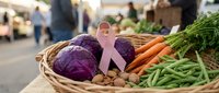 Fresh organic vegetables in wicker basket with pink awareness ribbon at farmers market. Purple cabbage, carrots, green beans for healthy eating and cancer support.
