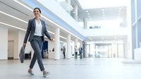 Businesswoman in formal attire walking through modern office lobby with tablet, representing corporate professional, business travel, workplace mobility, and contemporary work environment.
