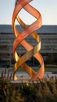 Modern twisted copper and brass spiral sculpture with intertwining ribbons in front of contemporary office building, suitable for corporate architecture and public art projects.