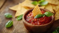Fresh homemade salsa with tomatoes, onions and herbs served in rustic wooden bowl with crispy corn tortilla chips and basil leaves on wooden table background.