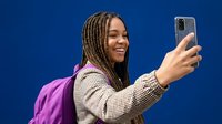 Young African American woman with braids taking selfie with smartphone against blue background. Student with purple backpack smiling at phone camera.