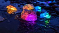 Glowing colorful rocks scattered on wet dark surface with vibrant pink, blue, orange and green illumination creating magical atmosphere in night setting.