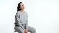 Young Asian woman in gray sweatsuit sitting relaxed with eyes closed, expressing peace and mindfulness on white background for wellness and lifestyle content.