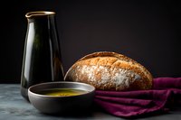 Rustic sourdough bread loaf with olive oil in ceramic bowl and dark bottle against black background, moody food photography on textured surface.