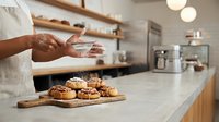 Chef with medium skin tone sprinkling flour over freshly baked croissants on wooden board in modern kitchen. Ideal for bakery, culinary, and food service marketing materials.