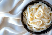 Fresh rice noodles in black ceramic bowl on white textile background, traditional Asian cuisine ingredient photographed from above with soft lighting.