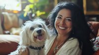 Hispanic woman with long dark hair smiling warmly while sitting on couch with small white fluffy dog. Natural lighting creates cozy home atmosphere. Pet companionship lifestyle.