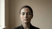 Young Caucasian woman with decorative floral face mask, eyes closed, serene expression, wearing dark robe, minimal background, skincare and wellness concept.
