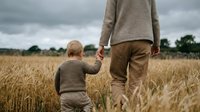 Father and young son walking hand in hand through golden wheat field on cloudy day. Family bonding moment in rural countryside during harvest season.