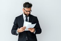 Bearded businessman in black suit and glasses reading documents against light background. Professional male executive reviewing paperwork in corporate setting.