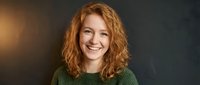 Cheerful young Caucasian woman with curly red hair smiling confidently against dark textured wall, wearing green sweater, studio portrait.