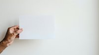 Close-up of a hand with medium-deep skin tone holding a blank white card or paper against a neutral background, suitable for mockups, advertising, and promotional designs.