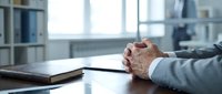 Businessman sitting at office desk with clasped hands next to an old book, conveying focus, strategy, and professional confidence.