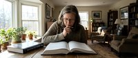 Middle-aged Caucasian woman with long gray hair reading book at wooden table in cozy home library with natural light from windows and bookshelves.