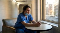 Tired healthcare worker in blue scrubs sitting alone in hospital break room during shift, looking exhausted and contemplative by window.