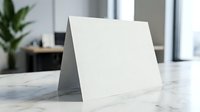 Blank white folded card standing on marble surface in modern office interior with plant background for branding mockup and presentation design projects.