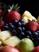 Fresh mixed fruit platter with juicy strawberries, dark purple grapes, golden pineapple chunks and green apples arranged in appetizing composition on dark background.