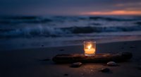 Glowing candle in glass holder on driftwood at twilight beach with ocean waves and colorful sunset sky creating peaceful romantic atmosphere for meditation.